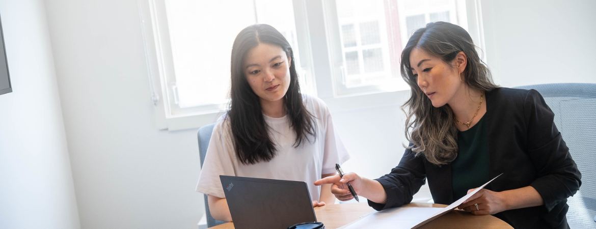 Two professionals sitting at a round table and looking at a laptop, with one of them pointing at the laptop.