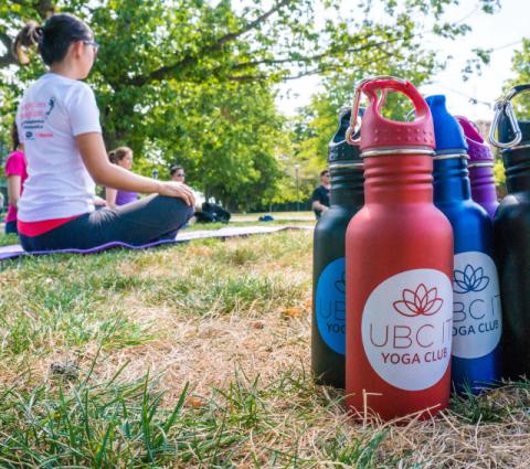 Colourful, reusable water bottles from the the UBC Yoga fund in the foreground, A group meditates in the background on the grass