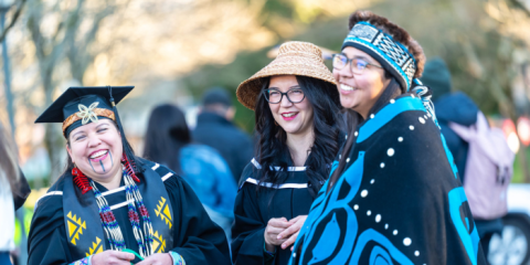 Three individuals dressed in Indigenous ceremonial garb talking and laughing outdoors.