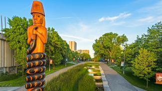 A large wooden statue of a figure holding a snake stands beside a pathway and a water feature. The path is lined with trees and banners, leading toward buildings in the background under a lightly clouded sky.